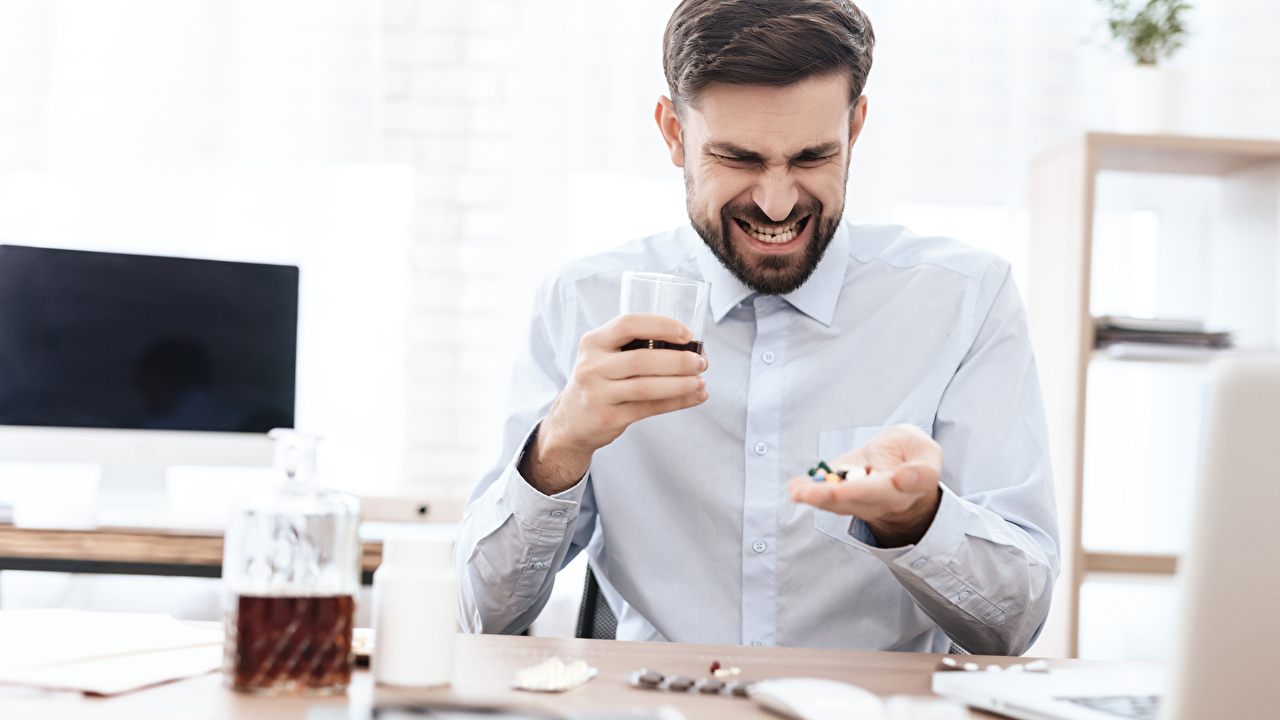 A man with pills in his hands sits at a table and suffers. A man with a beard sits with alcohol in his hands and holds a pill. A man with pills in his hands sits at a table and suffers. A man with a beard sits with alcohol in his hands and holds a pill.