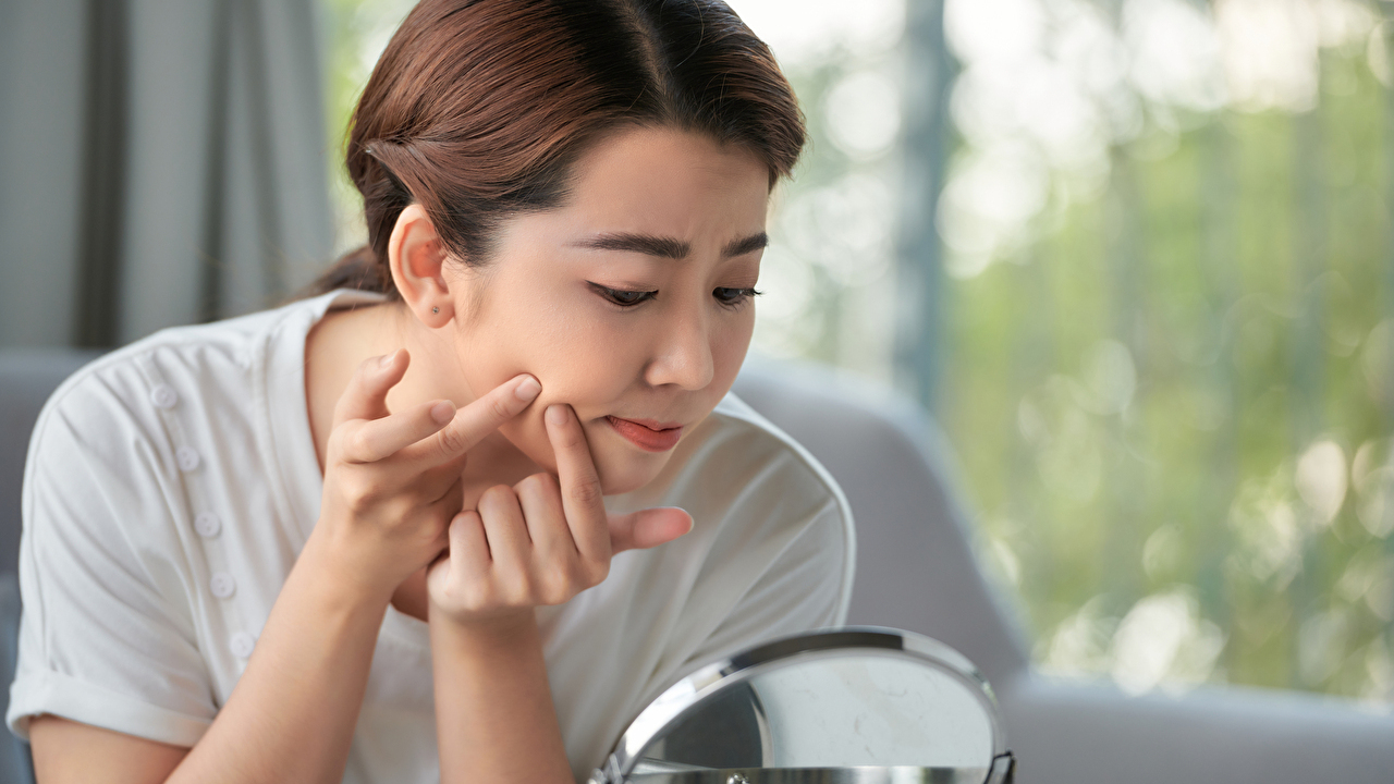 Girl looking at mirror and popping a pimple at home.